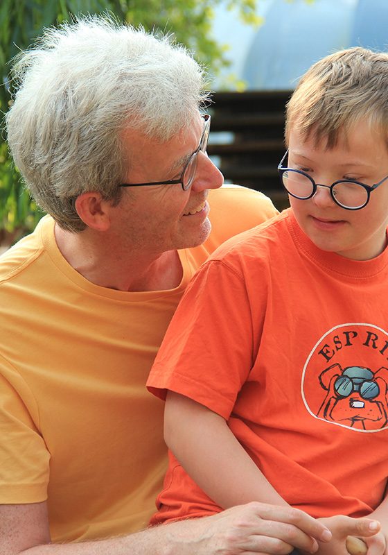 Ein Mann mit gelbem T-Shirt hat einen Jungen mit Down-Syndrom auf dem Schoß. Der Junge trägt ein oranges T-Shirt. Der Mann lächelt den Jungen an und spricht. Der Junge scheint aufmerksam zuzuhören.