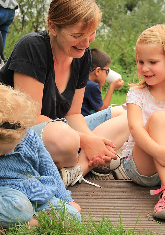 Eine Frau sitzt im Schneidersitz auf Brettern auf einer Wiese. Sie schaut nach unten und lacht. Neben ihr sitzt ein blondes, lachendes Mädchen mit angezogenen Knien. Die beiden scheinen sich zu unterhalten. Sommerliche, entspannte Stimmung.
