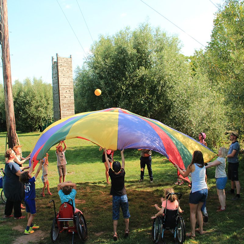 Mehrere Kinder und Erwachsene stehen im Kreis auf einer Wiese. Fast alle halten ein buntes Schwungtuch hoch in die Luft. Zwei Kinder sitzen in einem Rollstuhl. Alle haben Spaß zusammen.