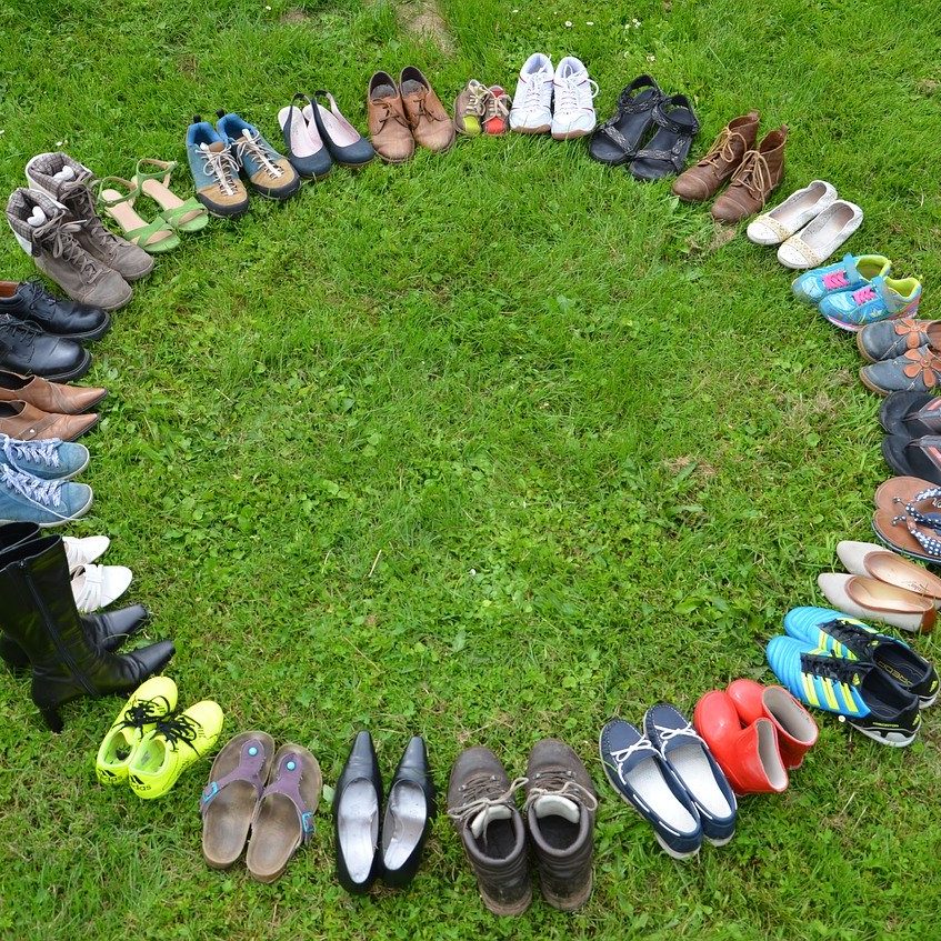 In grünem Gras sind etwa fünfzig Schuhe paarweise in einem Kreis aufgestellt. Die Schuhe sind bunt, haben unterschiedliche Größen und Formen. Es sieht so aus, als würden sie sich austauschen wollen.