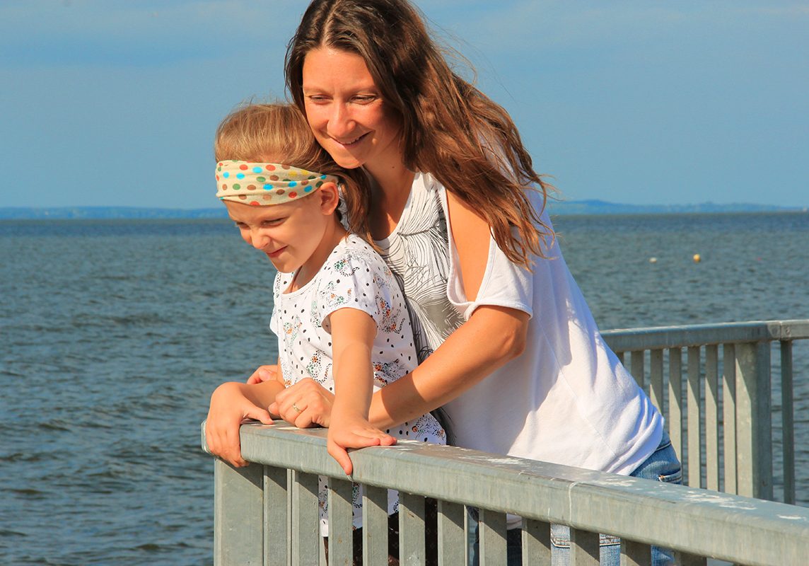 Ein blondes Mädchen mit buntem Stirnband steht auf einer Seebrücke. Sie hält sich am Geländer fest und schaut nach unten. Von hinten umarmt sie eine junge Frau mit langen Haaren. Im Hintergrund sieht man das Meer und den Horizont. Ferienstimmung.